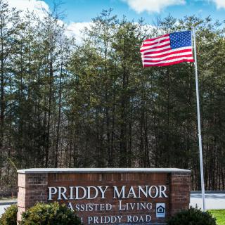 A sign for Priddy Manor Assisted Living with an American flag waving in the background.