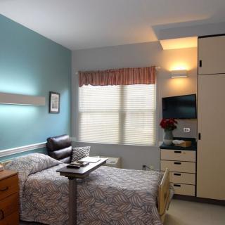 A neatly arranged hospital room with a single bed, TV, and furniture beneath the window.