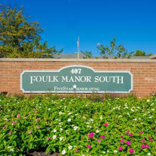 A sign reading 'Foulk Manor South' is mounted on a brick wall, surrounded by a lush garden with various flowers and set against a clear blue sky.