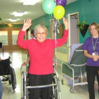 An elderly woman raises her hands in celebration, standing with the help of a walker in a healthcare facility, surrounded by balloons and supportive staff.