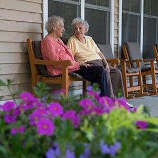 Two elderly women sitting on a porch having a conversation, with vibrant purple flowers in the foreground.