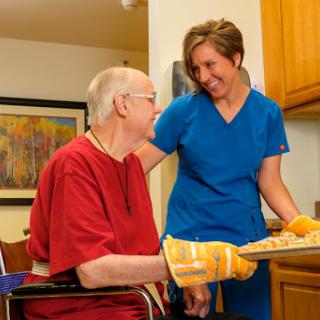 A caregiver assists an elderly person in a kitchen setting, both sharing a moment of joy.