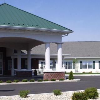 A large building with a covered entrance, featuring white columns and a green roof, surrounded by landscaping and a parking lot.