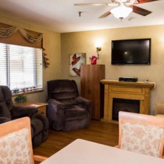 A cozy living room with recliner chairs, a television mounted above a fireplace, and a ceiling fan overhead. The room is well-lit by natural light coming through the window.