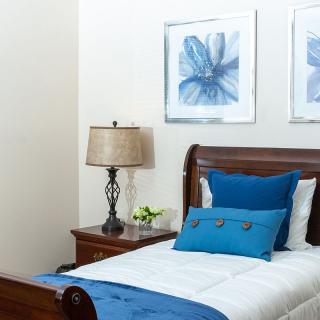 A neatly arranged bedroom with a single bed featuring blue decorative pillows and a runner, accompanied by a wooden nightstand with a lamp and floral artwork on the walls.