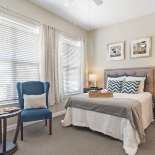 A cozy bedroom featuring a neatly made bed with decorative pillows, a blue armchair, and soft natural light streaming through large windows.