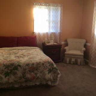 A cozy bedroom with a floral bedspread, two windows with curtains, a nightstand, and an upholstered chair.