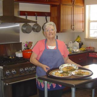 A smiling woman stands in a kitchen, holding a tray of food.