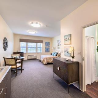 A well-lit bedroom with modern furnishings and decor, featuring a bed, dresser, seating area, and an adjacent bathroom.