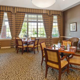 A cozy dining room with tables and chairs arranged on a patterned carpet, featuring large windows and elegant curtains.