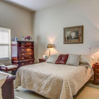 A cozy bedroom with a neatly made bed, adorned with pillows and a quilt, flanked by lamps on side tables. The room features elegant wooden furniture and wall art, creating a warm and inviting atmosphere.