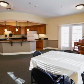A spacious dining area and kitchen with wooden cabinetry, a white table, chairs, and a piano by the window.