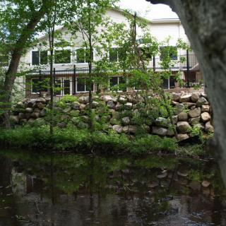 A picturesque view of a building by a river, surrounded by trees and a stone wall.