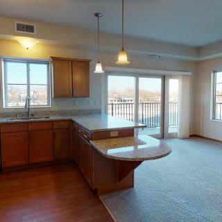 A modern open-plan kitchen with wooden cabinets and a large island countertop, featuring pendant lighting and ample natural light from windows and a sliding door.