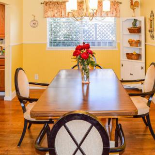 A bright dining room featuring a wooden table set with a vase of red flowers, surrounded by chairs, under a chandelier.