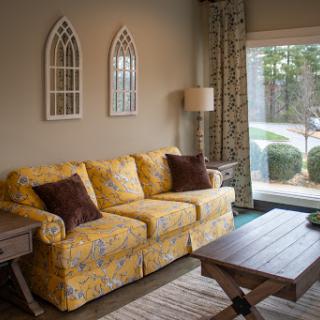 A cozy living room featuring a yellow patterned couch, wooden coffee table, and decorative window panels.