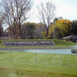 A scenic view featuring a large sign beside a calm water body, surrounded by lush grass and trees.