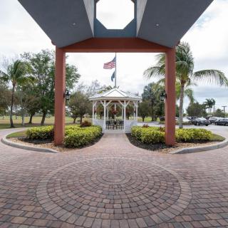 A scenic gazebo with a flagpole surrounded by palm trees and lush landscaping, viewed through a paved pathway.