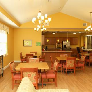 A cozy dining area with warm yellow walls, wooden tables and chairs, and a view into a kitchen.