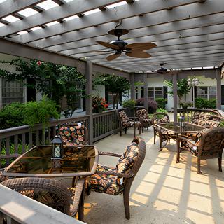 A cozy outdoor patio area featuring wicker furniture under a pergola, with lush greenery and a ceiling fan for comfort.