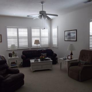 A cozy living room with sofas and armchairs arranged around a coffee table, featuring window shutters and a ceiling fan.