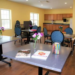 A bright community room with tables, chairs, and a small kitchen area.