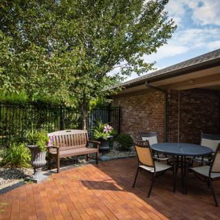 A cozy patio with a bench and table set under a tree, surrounded by brick walls on a sunny day.