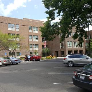 A view of a brick building surrounded by a parking lot with several vehicles and trees under a cloudy sky.