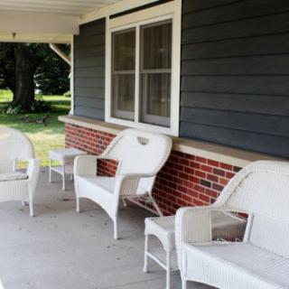 A cozy porch with white wicker furniture in front of a brick and dark siding house.