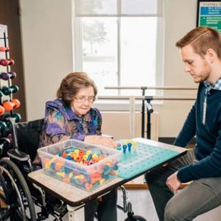 An elderly woman in a wheelchair engages in rehabilitation exercises with the assistance of a caregiver.