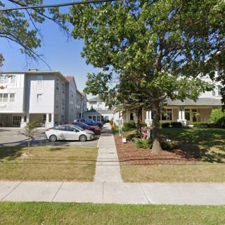 A suburban scene featuring apartment buildings with a driveway, parked cars, and surrounding greenery on a sunny day.