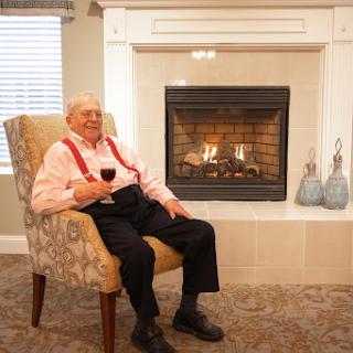 An elderly man enjoying a glass of wine by the fireplace in a cozy living space.