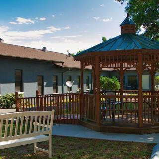 A peaceful outdoor area featuring a wooden gazebo and a bench, surrounded by greenery and adjacent to a building under a sunny sky.