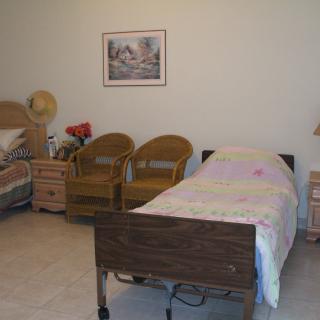 A cozy room featuring two hospital beds, wicker chairs, and a painting on the wall.