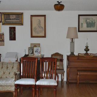 A vintage-style living room with various furniture pieces, including a sofa, chairs, a lamp, and a piano, complemented by artwork on the walls.