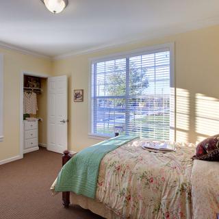 A cozy bedroom with natural light streaming through a large window, featuring a neatly made bed and soft carpeting.