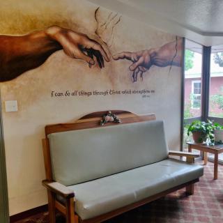 A mural featuring two hands reaching towards each other, inspired by a classic artwork, adorns the wall above a wooden bench in a cozy indoor space with a window and potted plant.