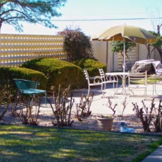 A sunlit patio area with outdoor furniture and an umbrella, surrounded by neatly trimmed bushes and a garden.