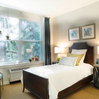 A cozy bedroom with a single bed, decorated with white bedding and accent pillows, featuring artwork on the wall and a large window allowing natural light in.