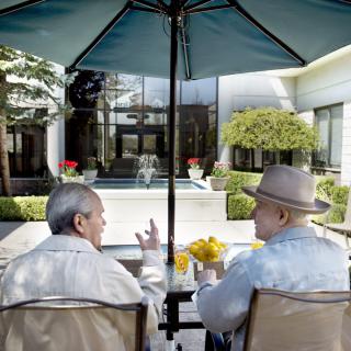 Two elderly individuals sitting under an umbrella, engaged in conversation on a patio.
