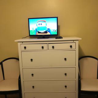 A television sits atop a white dresser, flanked by two chairs in a simple room setting.