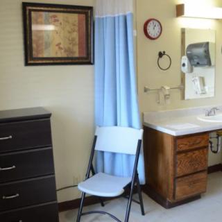 A small room featuring a folding chair, dresser, sink area with a mirror, and a curtain.