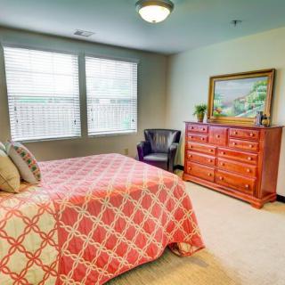 A cozy bedroom featuring a bed with a patterned comforter, a wooden dresser with a painting on top, and a chair in the corner.