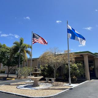 A view of a building with an American flag and a Finnish flag flying in front, accompanied by palm trees and a fountain.