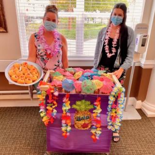 Two people in masks stand by a colorful, Hawaiian-themed cart with snacks.