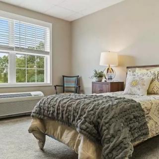A cozy bedroom with a large window, featuring a neatly made bed, bedside table with lamp, and elegant decor.