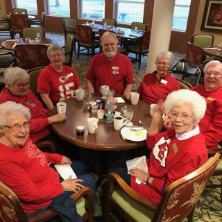 A group of seniors wearing matching red shirts gather around a table in a dining area.