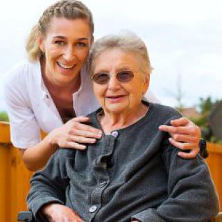 A caregiver smiling alongside an elderly person in a wheelchair outdoors.