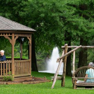 An outdoor scene featuring a gazebo and a swing with two people, set against a backdrop of trees and a fountain.