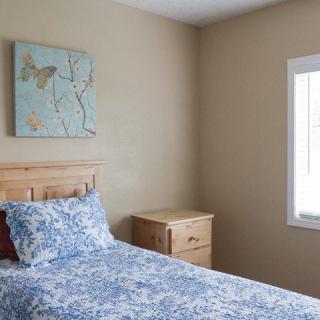 A cozy bedroom with a single bed featuring blue patterned bedding, a wooden headboard, and a light wood nightstand. A piece of wall art hangs above the bed, and natural light streams in through the window.